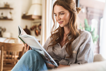 Positive optimistic redhead woman indoors sitting at home reading book or magazine.