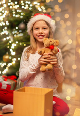 christmas, holidays and childhood concept - smiling girl in santa helper hat with gift box and teddy bear at home