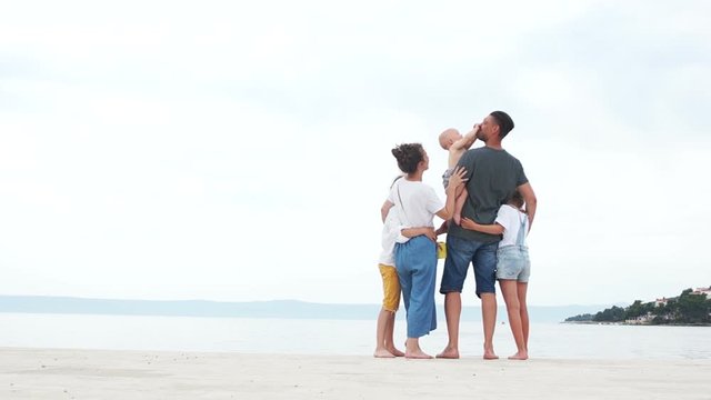 A large family is resting on the seashore. Parents and three children. The kid in his fathers arms reaches out to kiss mom and dad funny opening his mouth