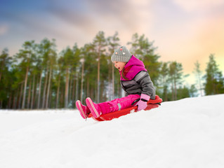 Fototapeta na wymiar childhood, sledging and season concept - happy little girl sliding down on sled outdoors over winter forest background