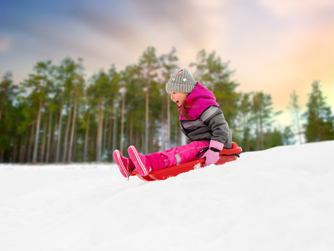 childhood, sledging and season concept - happy little girl sliding down on sled outdoors over winter forest background
