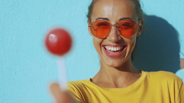 close-up of face of a young tanned laughing woman. cheerful smiling woman with red eyewear standing against blue background and joyfully licking a big red lollipop. Female bright make-up for lips
