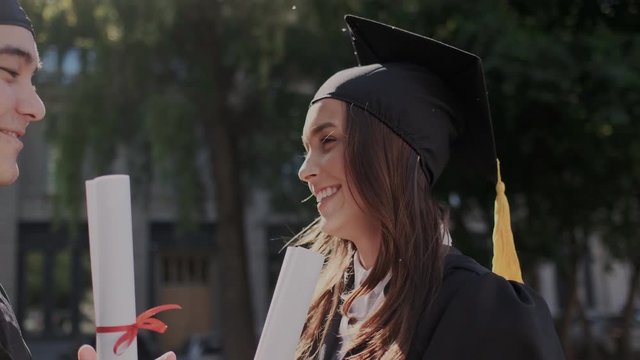Charming female graduate laughs and talks to her classmate friend. They dressed in traditional academic caps and mantle. Status diploma in hands. Concept of study and knowledge. Close-up, slow-motion.