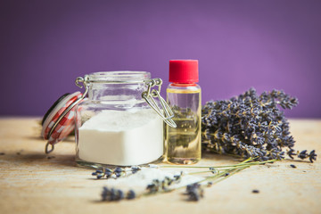 Olive oil and baking soda exfoliating scrub concept. Homemade natural beauty cosmetics. Jar with baking soda powder and bottle with olive oil, lavender bouquet on purple background.