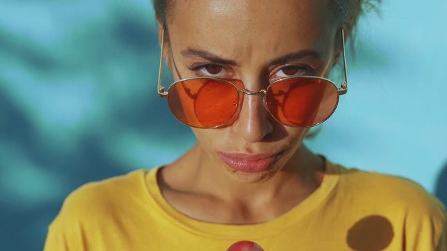 close-up of face of a young tanned emotional woman with big red lollipop. cheerful woman with red eyewear standing against blue background, licking lollipop and showing different emotions.