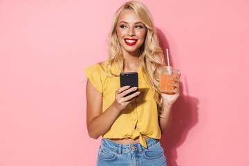 Smiling cheery young blonde woman using mobile phone drinking juice.