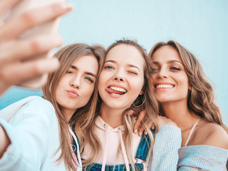 Three young smiling hipster women in summer clothes.Girls taking selfie self portrait photos on smartphone.Models posing in the street near wall.Female showing positive face emotions.Showing tongue