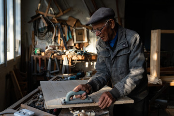 Very old master carpenter in glasses polishes wood in the home workshop, working hard