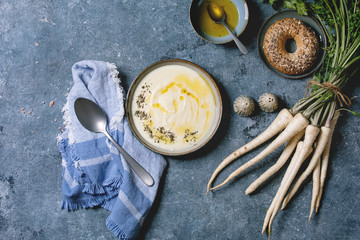 Parsnipcream soup in ceramic bowl with butter sauce, bagel bread, bundle of fresh parsnip and herbs over blue texture background. Flat lay, space - obrazy, fototapety, plakaty