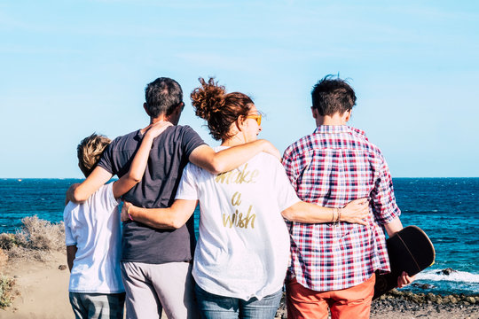 happy family together and hugged at the beach n front and looking at the sea - four persons - child and teenager - millennial with skate board in his arm