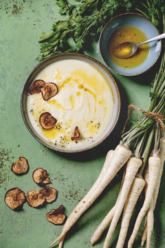 Parsnip cream soup in ceramic bowl with sun dried pears, butter sauce, bundle of fresh parsnip and herbs over green texture background. Flat lay, space