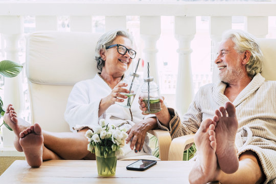 Couple Of Seniors Or Mature People In A Resort Spa In Their Hotel Or House Clinking With Their Cocktail Or Drink - Having Fun With Feet On The Table Looking Between They