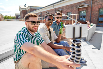 leisure, technology and people concept - happy male friends taking selfie by smartphone on tripod and drinking beer on street in summer