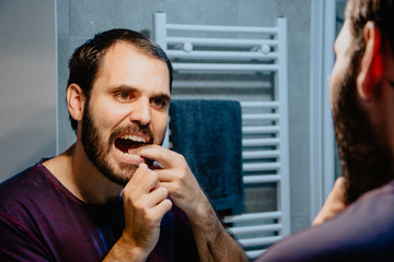Cleaning teeth with dental floss. Rear view of handsome young cleaning his teeth with dental floss and smiling while standing in front of the mirror