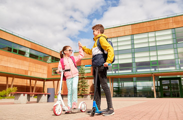 education, childhood and people concept - happy school children with backpacks and scooters making high five outdoors