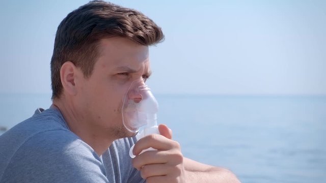 Portrait sick man uses nebulizer on seaside. Inhaling inhaler mask. Fibrosis cystic copd and treatment inhaler. Asthma pulmonary respiratory breath problem and cure. Painkiller sedative gas oxygen.