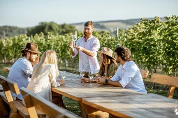Fototapeten Gruppe junger Leute, die Wein trinken und miteinander reden, während sie an einem sonnigen Abend am Esstisch im Freien auf dem Weinberg sitzen? © rh2010