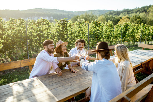 Group of a young people drinking wine and talking together while sitting at the dining table outdoors on the vineyard on a sunny evening