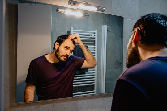 A worried young white man looks at himself in the mirror and inspects his premature receding hairline. Attractive Caucasian male adult in his 30s concerned about losing hair. Male pattern baldness