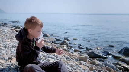 Sick child boy uses nebulizer sits on seaside throw stones. Inhaling inhaler mask. Fibrosis cystic copd and treatment. Asthma pulmonary respiratory breath problem cure. Painkiller sedative gas oxygen.