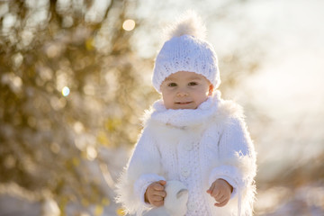 Baby playing with teddy in the snow, winter time. Little toddler boy in handmade white snowsuit,...