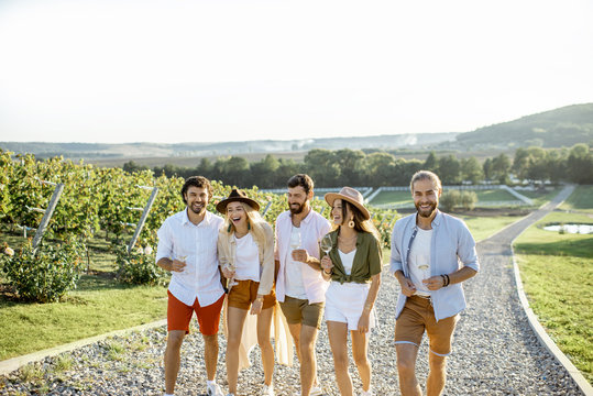 Group Of Young Friends Dressed Casually Hanging Out Together, Walking With Wine Glasses On The Vineyard On A Sunny Day