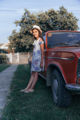 Full length portrait, beautiful young brunette little girl in dress and hat leaned on the red old antique car and looking at the caamera
