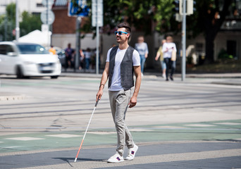 Young blind man with white cane walking across the street in city.
