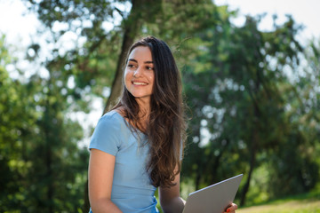 Obraz premium Young brunette student in a blue dress sitting with a laptop in a park. Distance learning