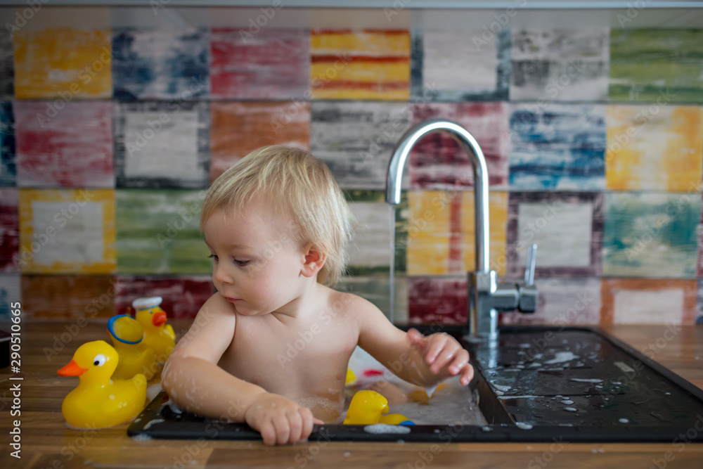 Poster Cute smiling baby taking bath in kitchen sink. Child playing with foam and soap bubbles in sunny kitchen with rubber ducks