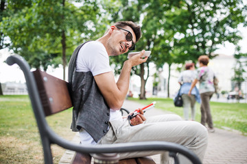 Fototapeta na wymiar Young blind man with smartphone sitting on bench in park in city.