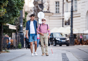 Young man and blind senior with white cane walking on pavement in city.