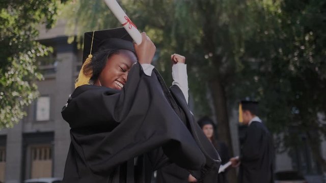 Close-up portrait of the African American happy young graduated woman. She having fun, dancing and making YES gesture. University and Graduates with on the background.