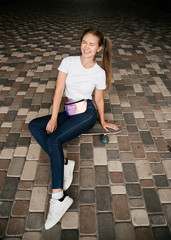 A happy young girl in white t-shirt and blue jeans is sitting on a skate board among the street.