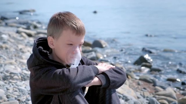 Portrait sick 8y child boy uses nebulizer sits on seaside. Inhaling inhaler mask. Fibrosis cystic copd and treatment. Asthma pulmonary respiratory breath problem cure. Painkiller sedative gas oxygen.