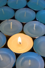 Closeup of tea lights with one being lit. A single warm orange flame between  candles in blue shadow. Christmas, mourning, condolence, memorial, funeral or cremation ceremony concept. Vertical image  