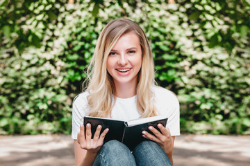 Young blonde girl reads a book and laughs in a park on a background of trees and bushes