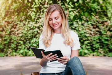Young blonde girl sits on a bench, reads a book and smiles in a park on a background of trees and bushes