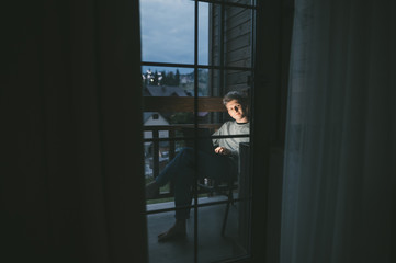 Positive young man sitting in chair in the evening on the balcony at home with a laptop on his lap, looking into the camera and smiling. Portrait of happy freelancer working on computer in apartment