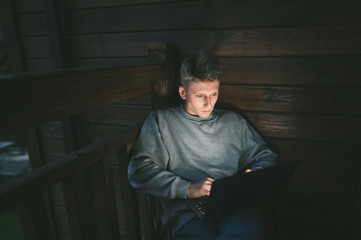 Portrait of handsome young man sitting on balcony in chair and using laptop, looking at screen with serious face. Guy works on a laptop in the evening in the apartment. Freelancer works at home.