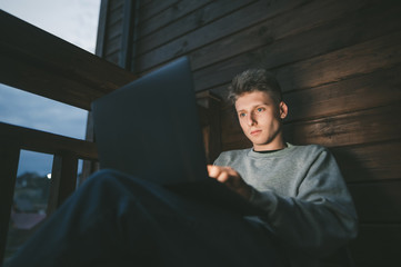 Cute guy sitting in the evening on a wooden balcony of a country house with a laptop on his lap and working, looking at the computer screen. Freelancer working in the evening on a laptop
