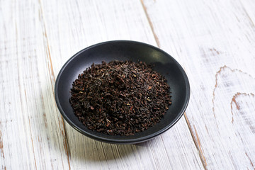 Black dry tea in a clay bowl on a light wooden background.