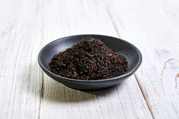 Black dry tea in a clay bowl on a light wooden background.