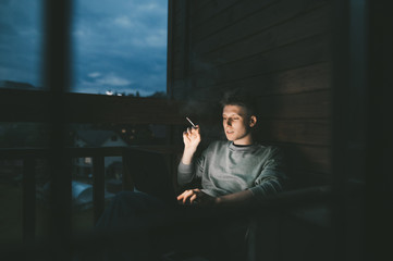 Happy young man sitting in chair in the evening on balcony in apartment, smoking and using laptop. Freelancer works on a laptop in a country house and smokes a cigar, looks at the window and smiles