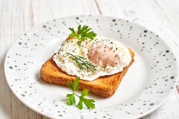 Poached egg, benedict on toasted white bread toast with salad and spices on a plate on a light wooden background.