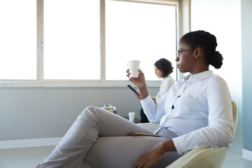 Fototapeta premium Office employees drinking coffee in office lounge. Young business woman sitting in armchair, holding and looking at disposal cup. Work balance concept