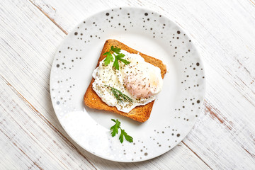 Poached egg, benedict on toasted white bread toast with salad and spices on a plate on a light wooden background.