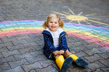 Happy little toddler girl in rubber boots with rainbow sun and clouds with rain painted with...