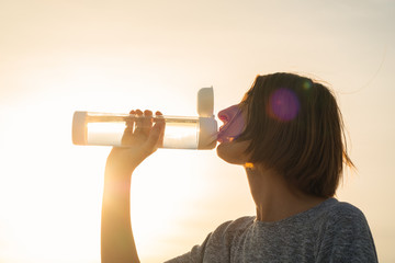 Young woman drinks water from a bottle on a sunset sky background. Portrait of a female with a reusable water bottle at sunset, thirst and rehydration concept