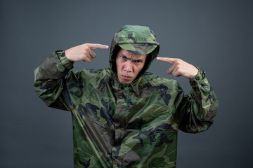 The young man wears a camouflage raincoat and shows different gestures. On a gray background in the...